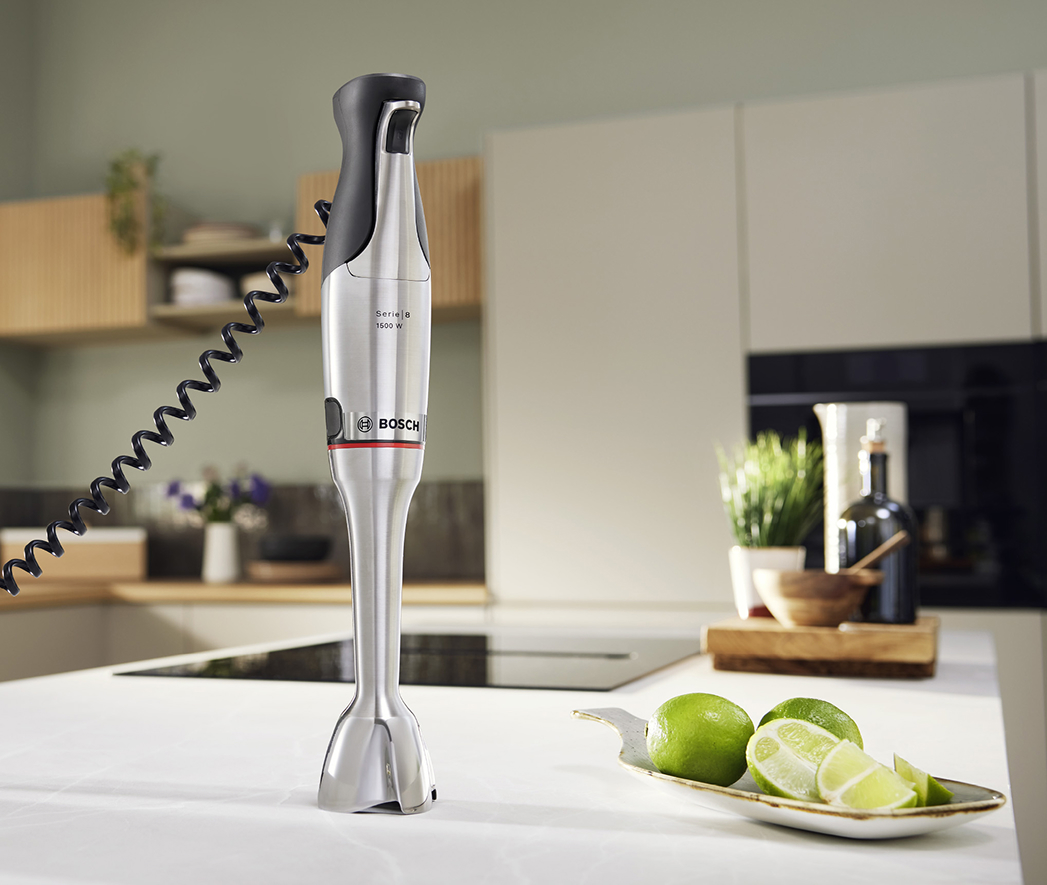 Pionowo ustawiony blender ręczny Bosch Serie 8 ErgoMaster 1500 W z obudową ze stali szczotkowanej i czarnym elastycznym kabel-spiralą. Urządzenie stoi na jasnym blacie nowoczesnej kuchni, obok leżą pokrojone limonki, a w prawym górnym rogu znajduje się niebieskie kółko z napisem 'x10'.